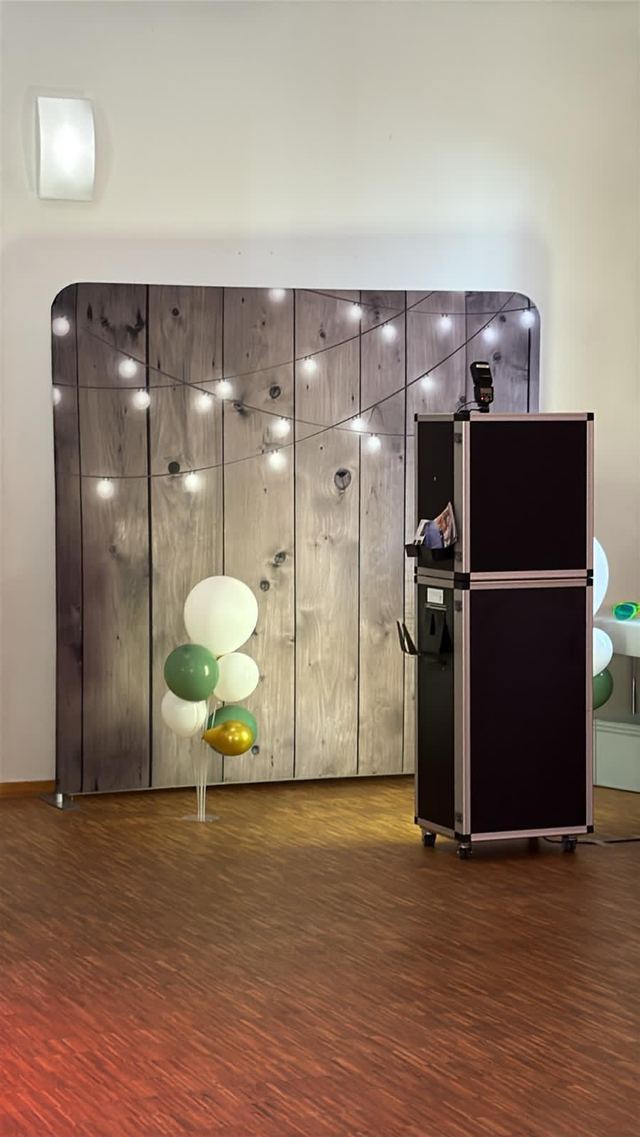 KOStBAR-Fotobox vor einer Holz-Fotowand mit Lichterkette und Luftballons